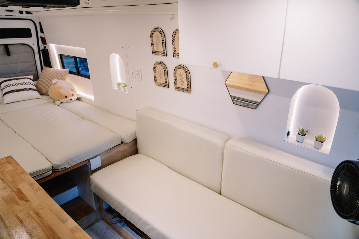 Cozy, modern RV rental interior featuring a bright white van space, cream cushions on bed and seating, minimalist decor, and soft lighting.