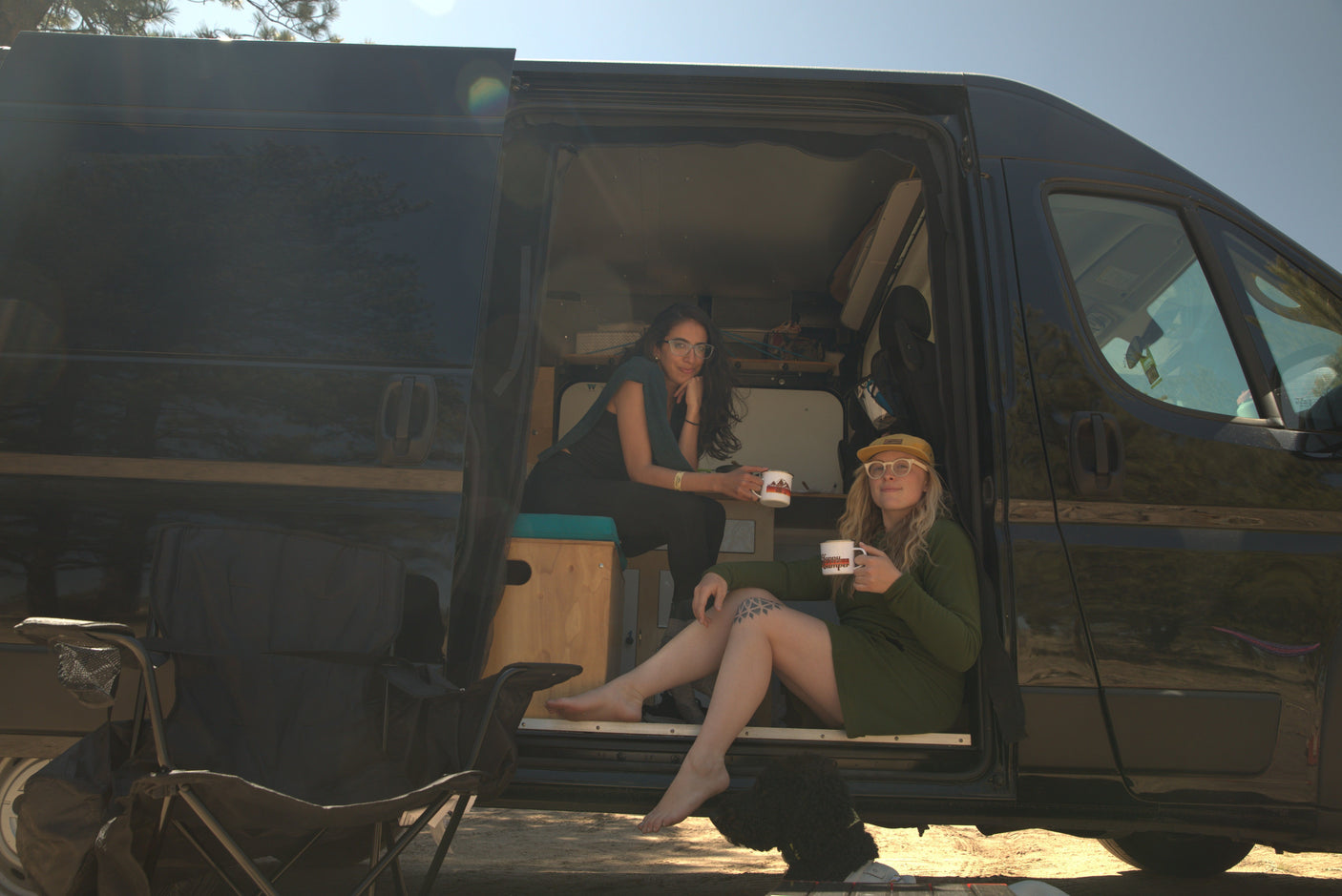 Two friends relaxing inside a black RV rental camper van, enjoying coffee mugs on a sunny outdoor day.