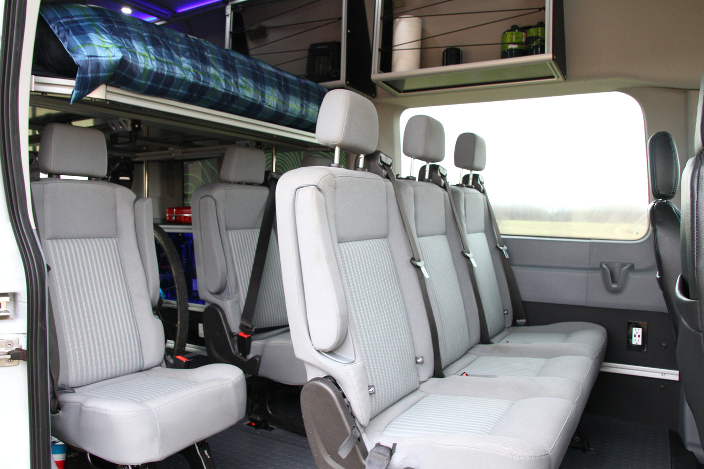 View of an RV rental's versatile interior, showing comfortable grey passenger seating, an overhead bunk, and storage.