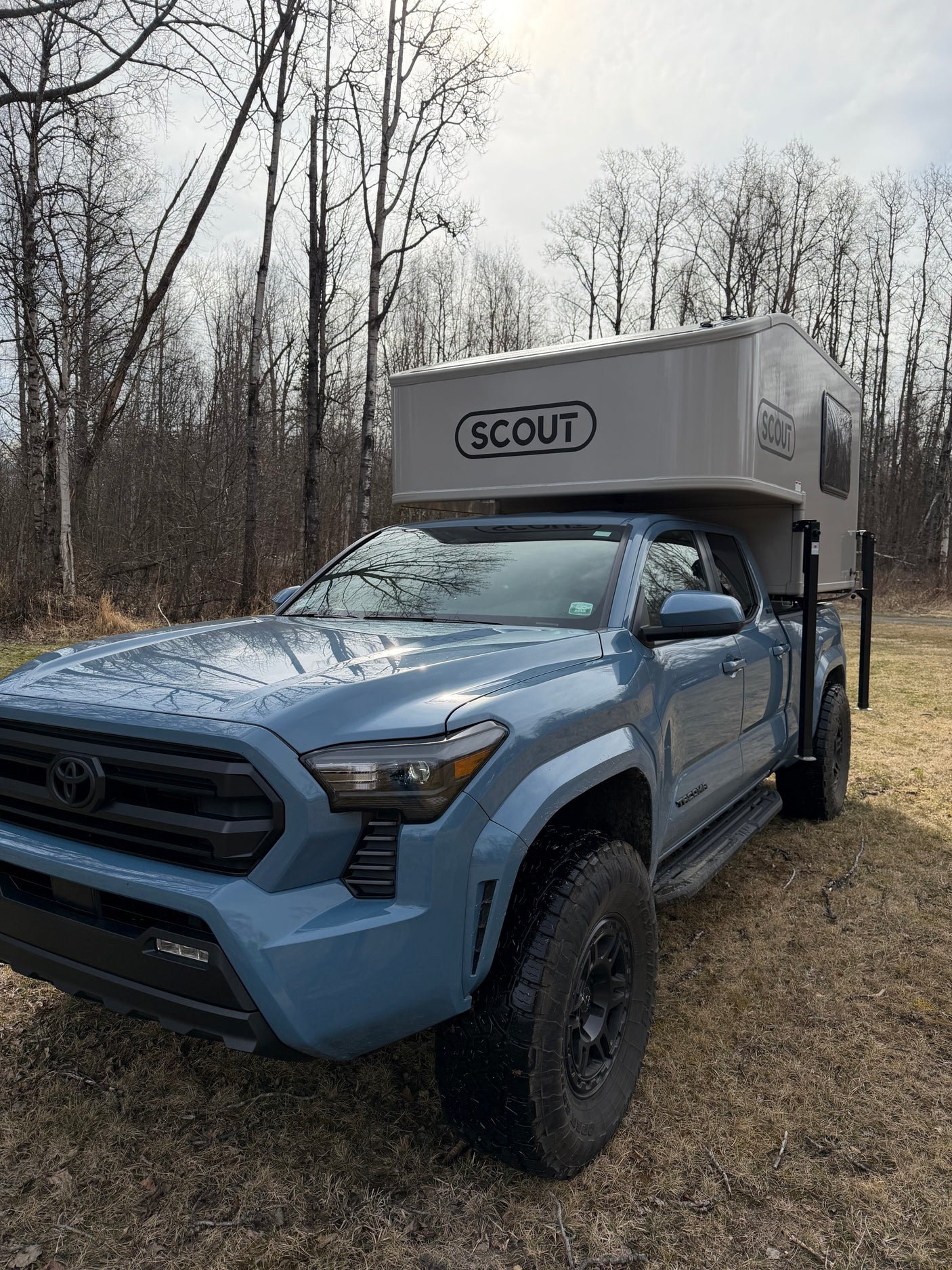 Blue Toyota Tacoma with a Scout truck camper, parked by bare trees. An excellent RV rental option for wilderness exploration.