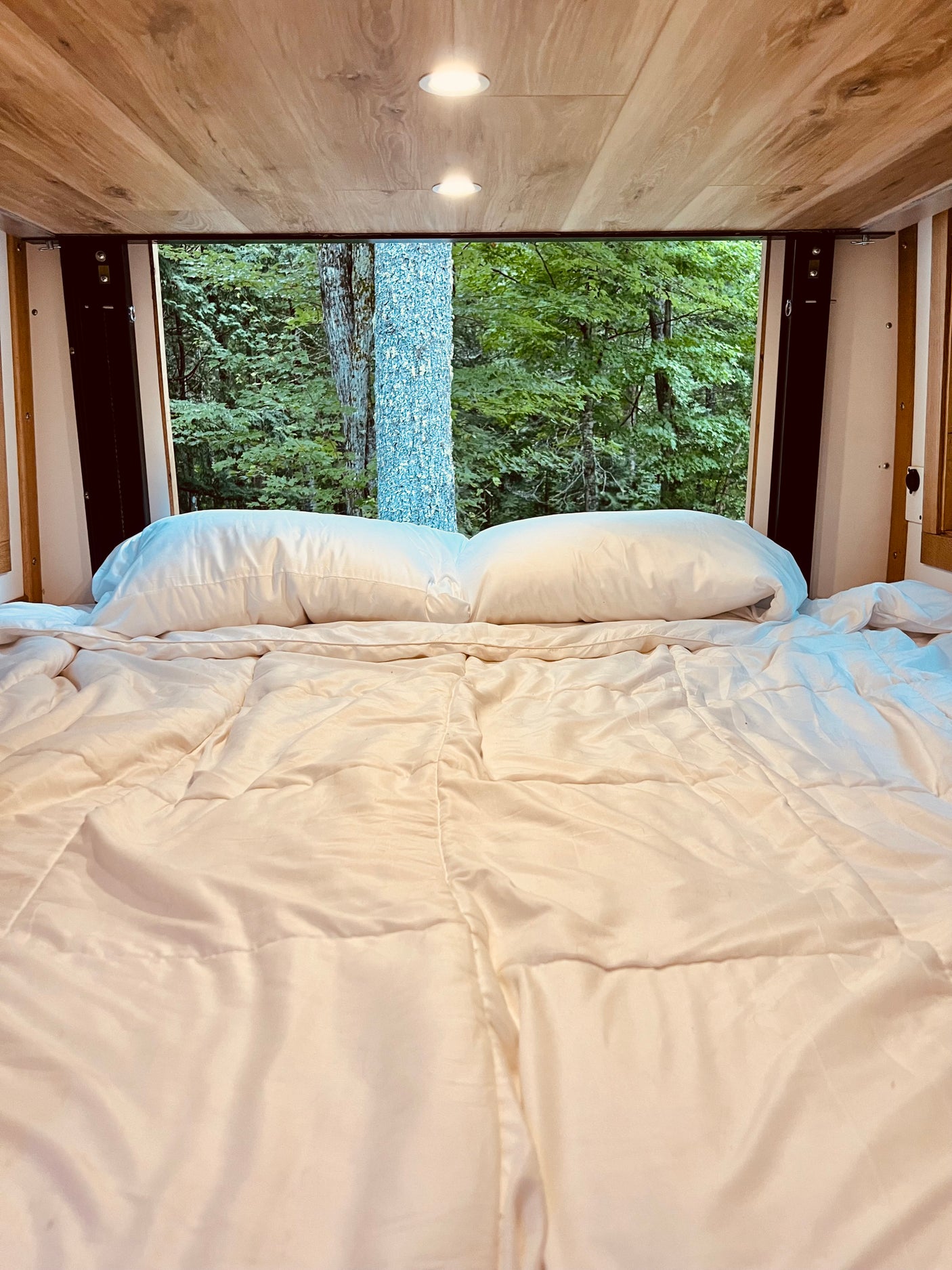 Enjoy a serene forest view from the cozy bed of this RV rental, featuring white bedding and a warm wooden ceiling with lights.