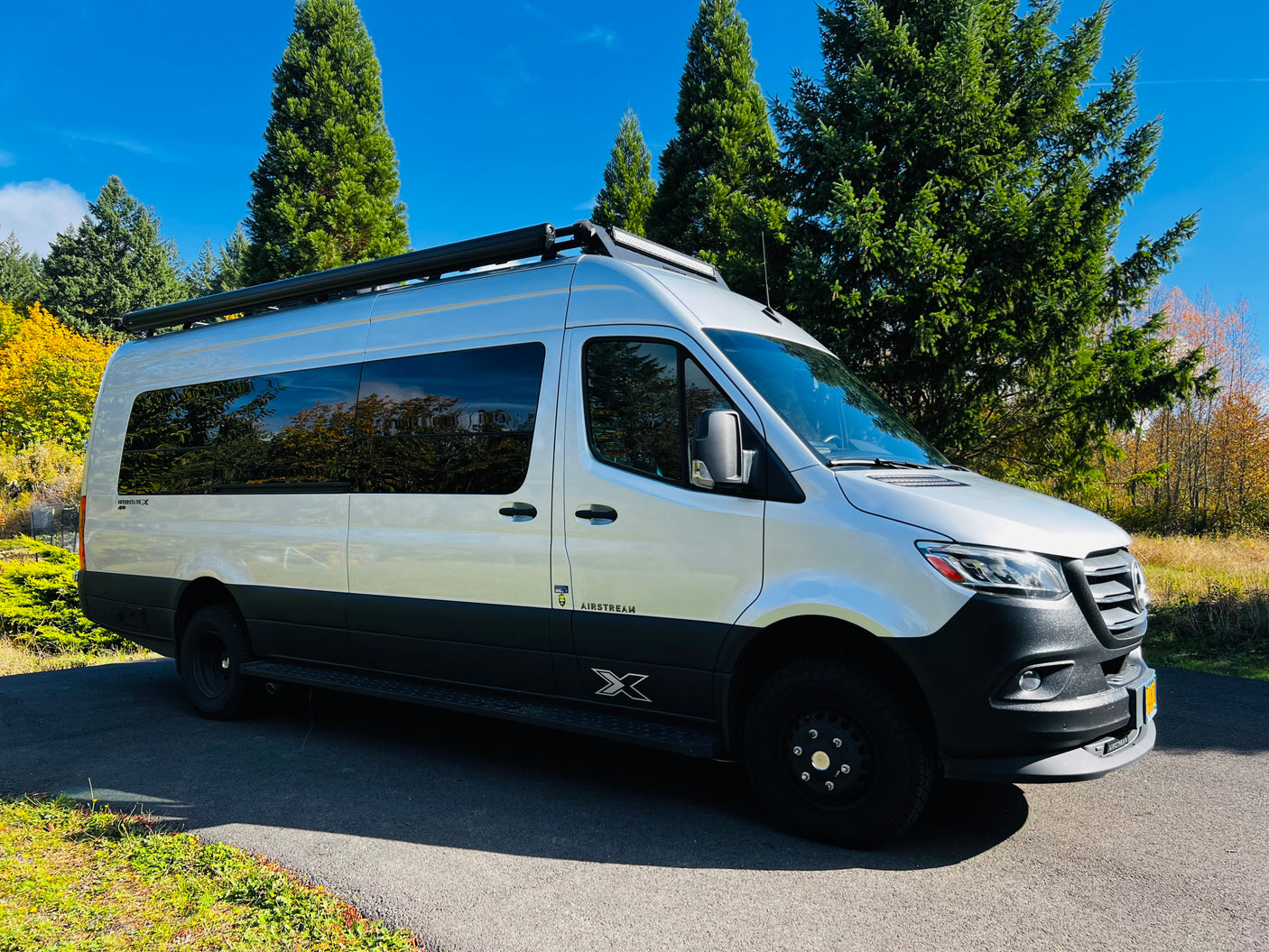 Silver Airstream Interstate X camper van, ideal for your next rv rental adventure, parked on a sunny day amidst tall green trees.