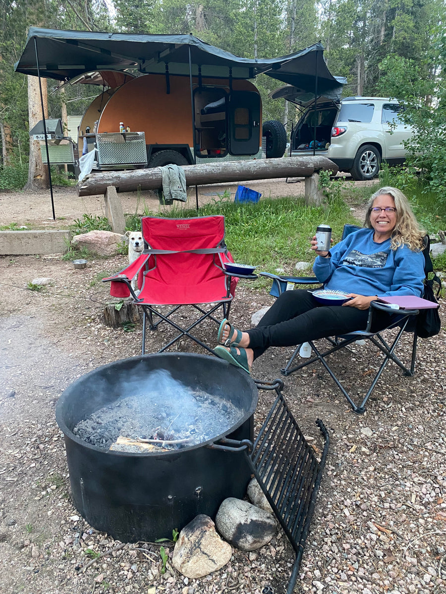 Enjoying a cozy RV rental campsite. Woman, dog, teardrop camper, & SUV relax by the fire.
