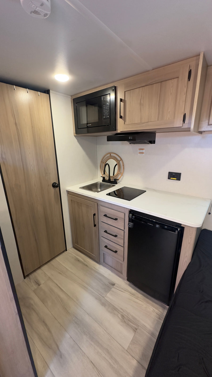 Modern RV rental kitchenette: light wood cabinets, black microwave, sink, cooktop, and compact fridge are visible.