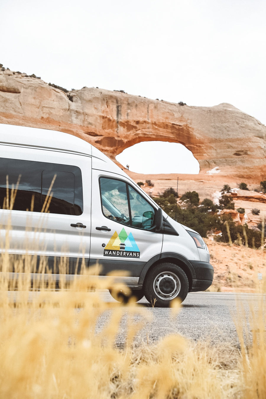 Silver RV rental van with 'Wandervans' logo by a desert road, facing a natural rock arch with distant figures.