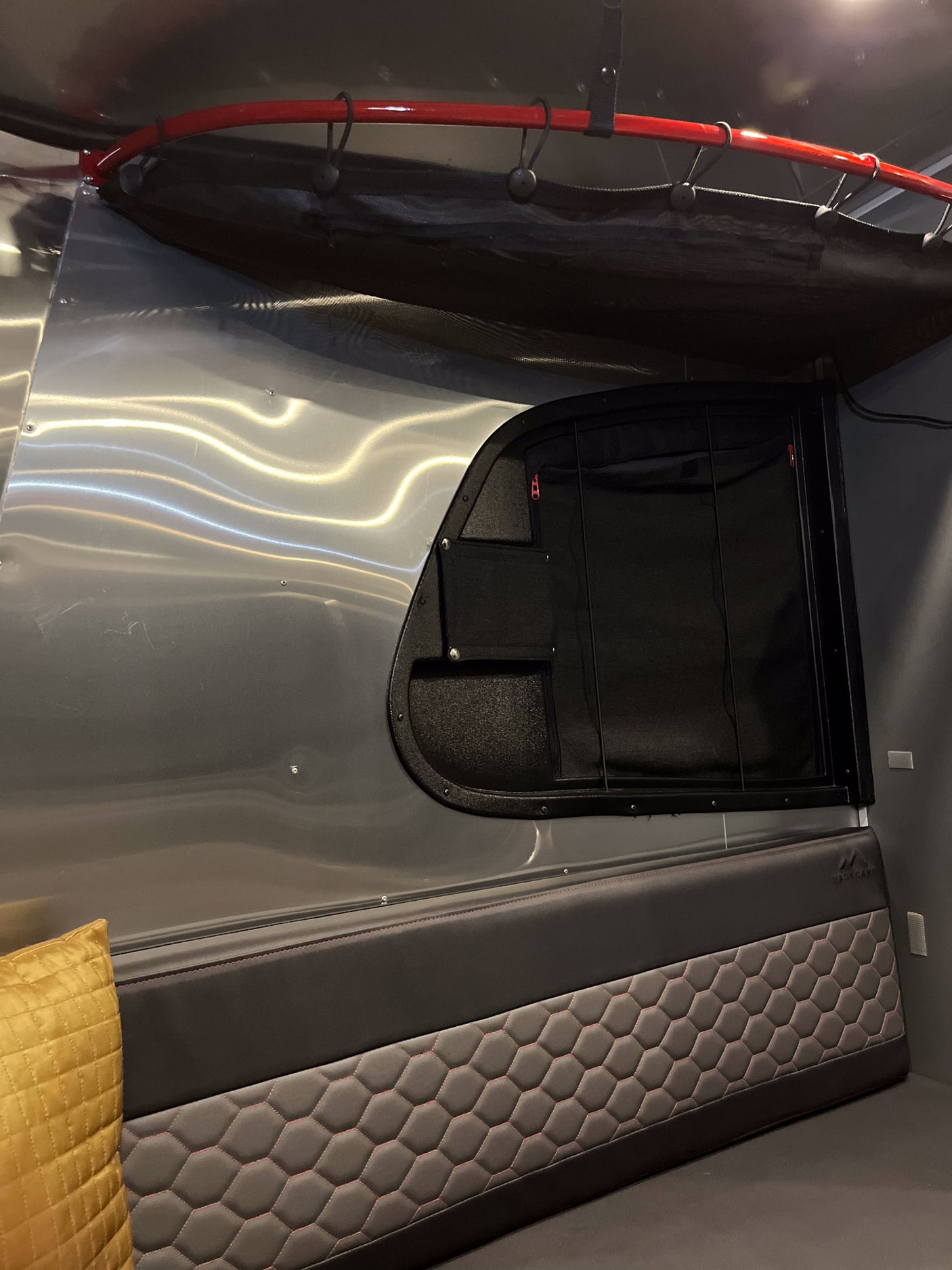 Inside this RV rental: a modern space with shiny walls, a red overhead bar, and hexagonal quilted seating with a yellow pillow.