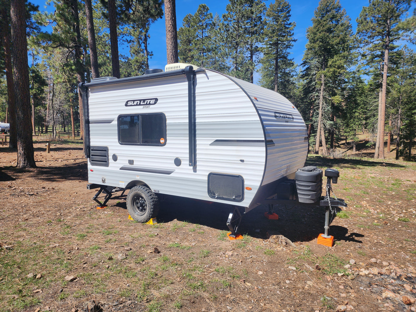 Sun Lite Sport travel trailer, ideal for RV rental, set up in a sunny forest campsite. White and gray, with orange stabilizing blocks.