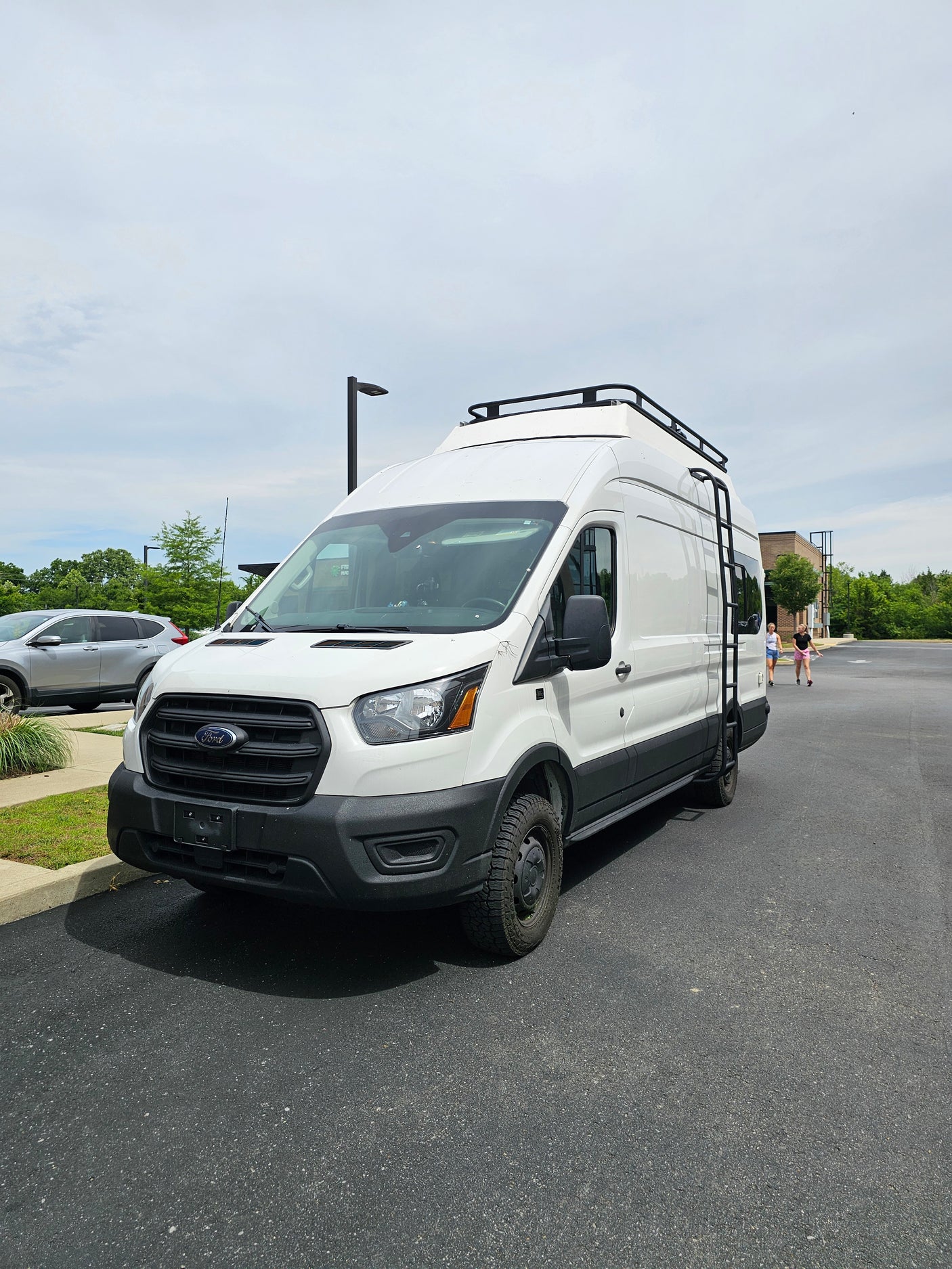 This white Ford Transit camper van, likely an RV rental, features a roof rack & side ladder.