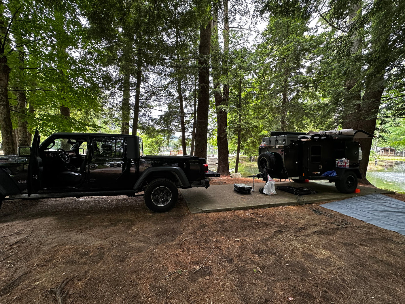 RV rental setup: A black Jeep Gladiator and an adventure trailer are parked on a concrete pad by a lake in a wooded campsite.