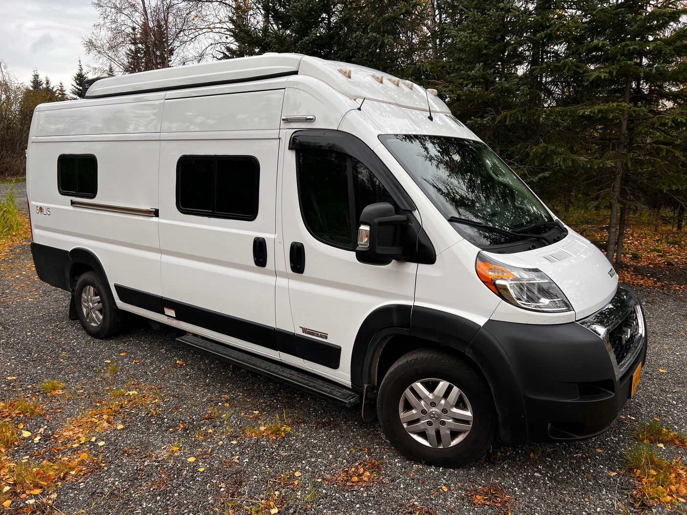 White Winnebago Solis RV rental van, ideal for nature travel. Parked on gravel among autumn trees.
