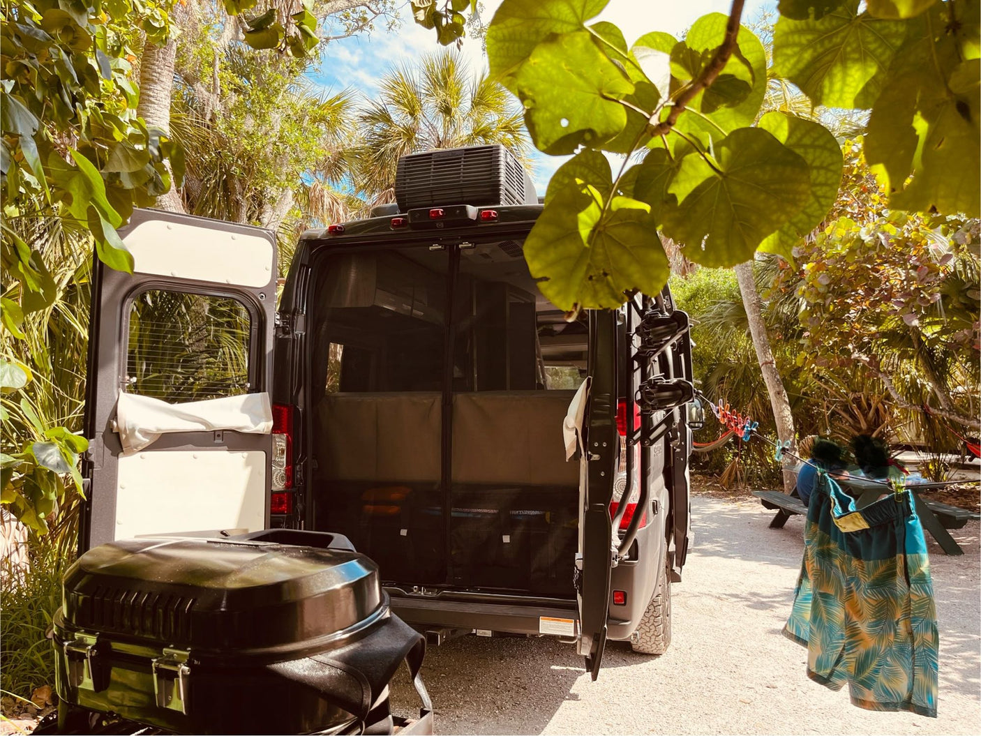 RV rental adventure: Black camper van parked at a tropical campsite with open doors, a grill, and swim trunks drying.
