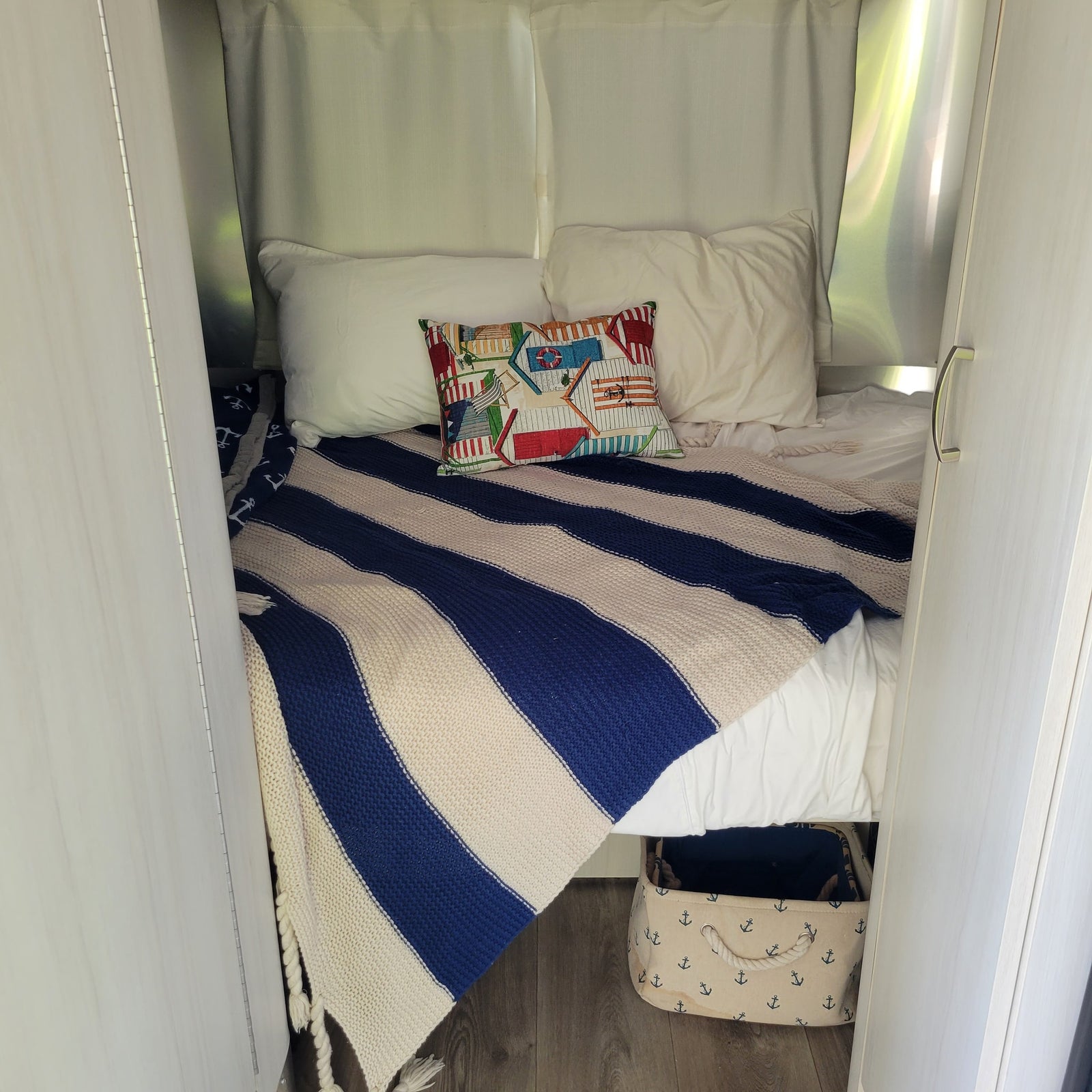 This RV rental's cozy bed features a blue striped blanket, beach house pillow, and anchor bin for storage.