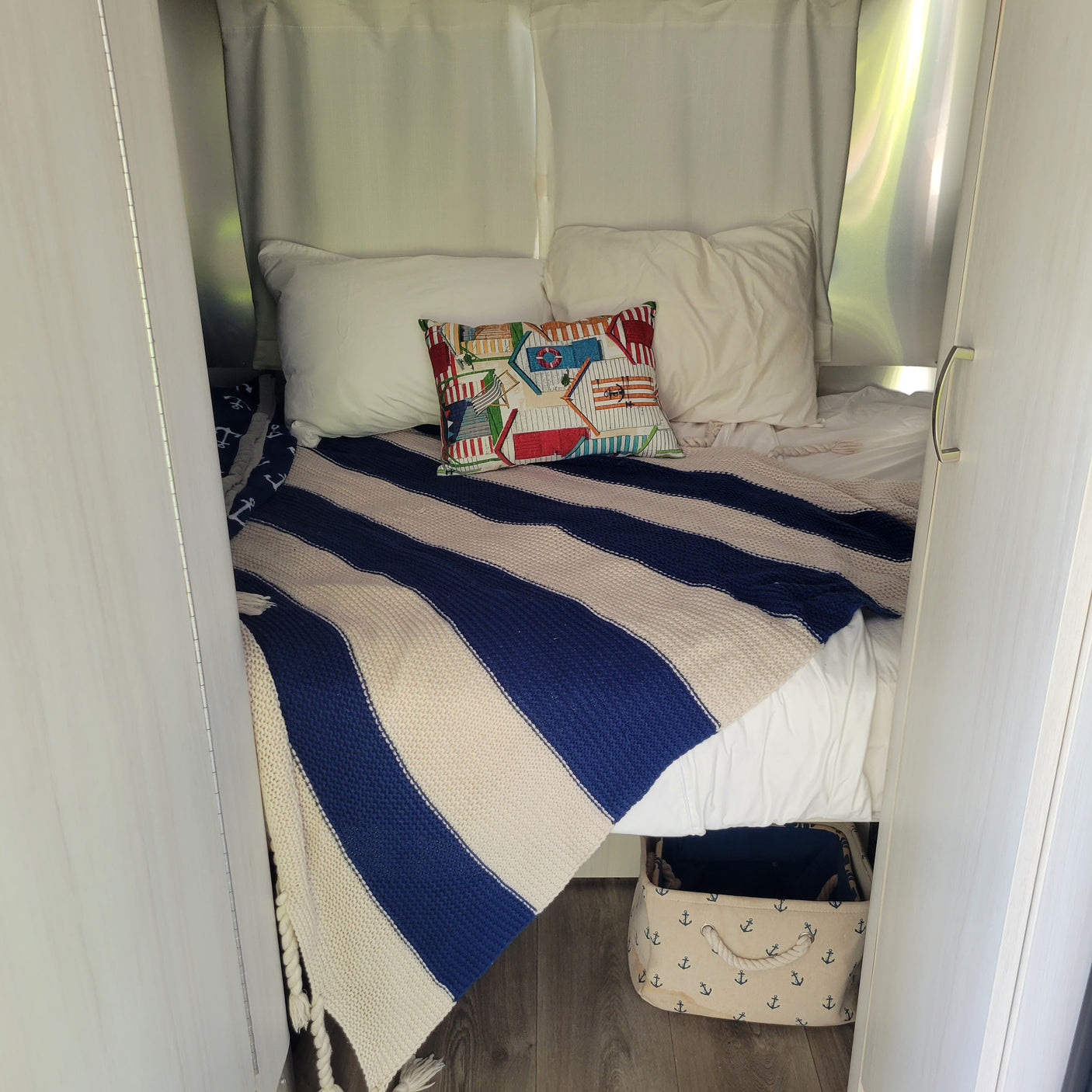 This RV rental's cozy bed features a blue striped blanket, beach house pillow, and anchor bin for storage.