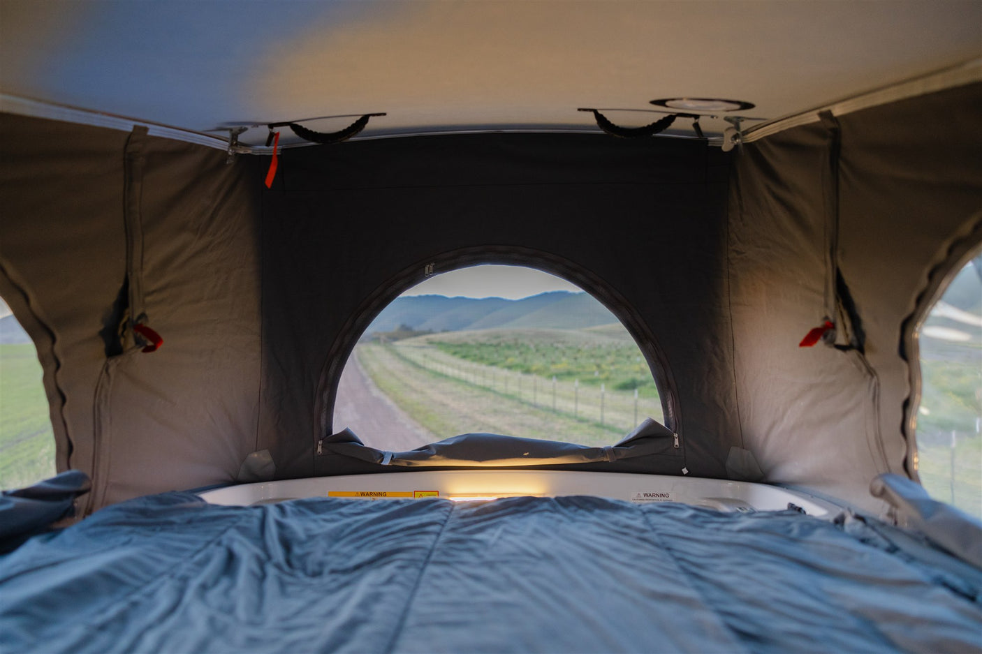 Inside a pop-top RV rental: a cozy bed with an arched window view of a road and rolling hills.