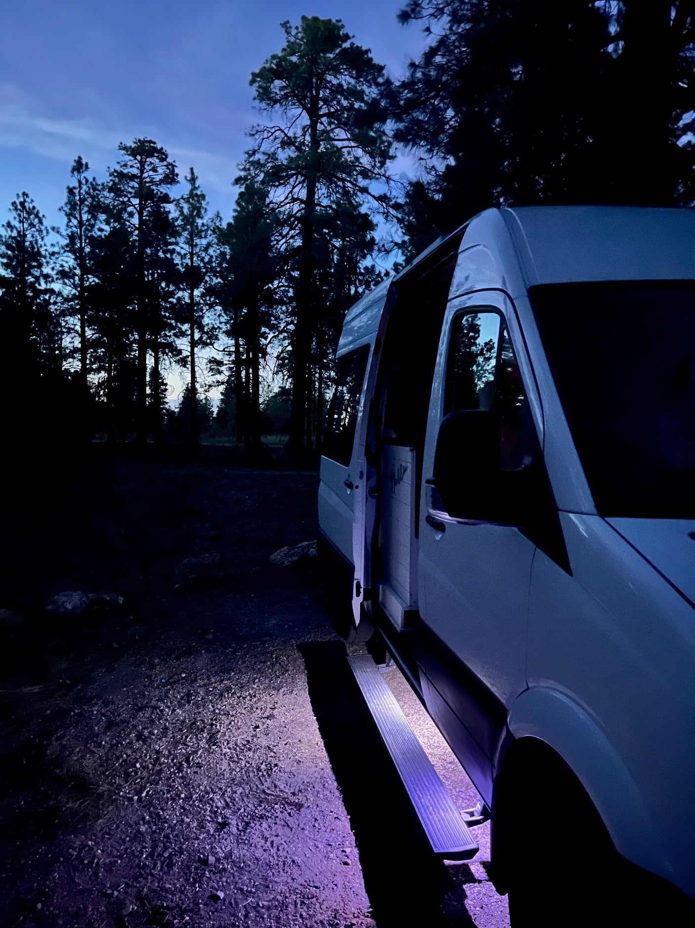 White **RV rental** van parked at night with open door and glowing purple step in a dim forest. Ready for adventure.