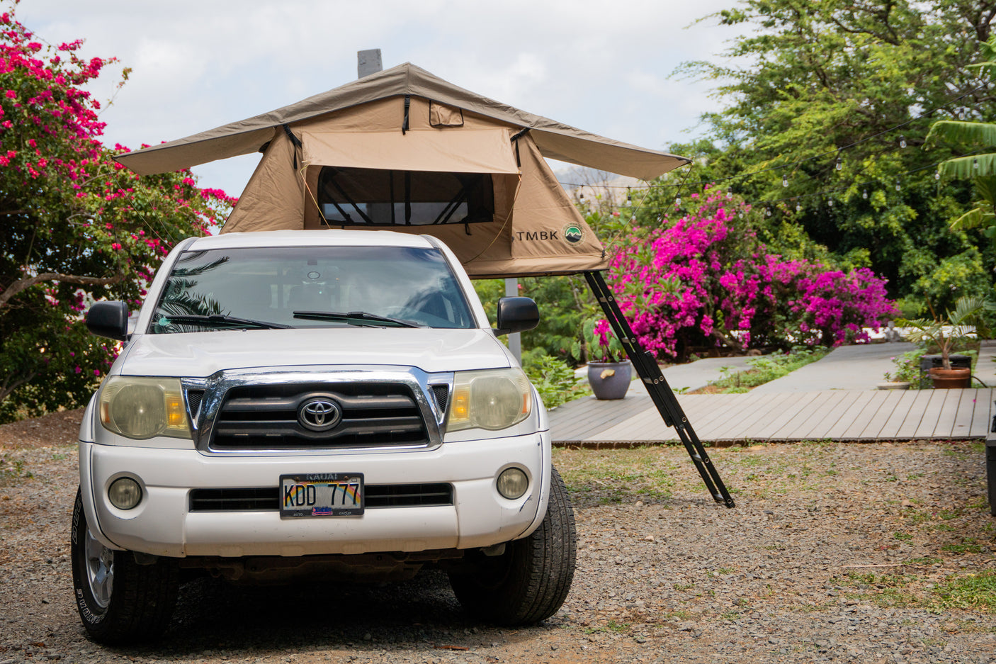 White pickup truck with a deployed rooftop tent, offering a unique RV rental for camping. Parked on gravel amidst lush pink flowers.