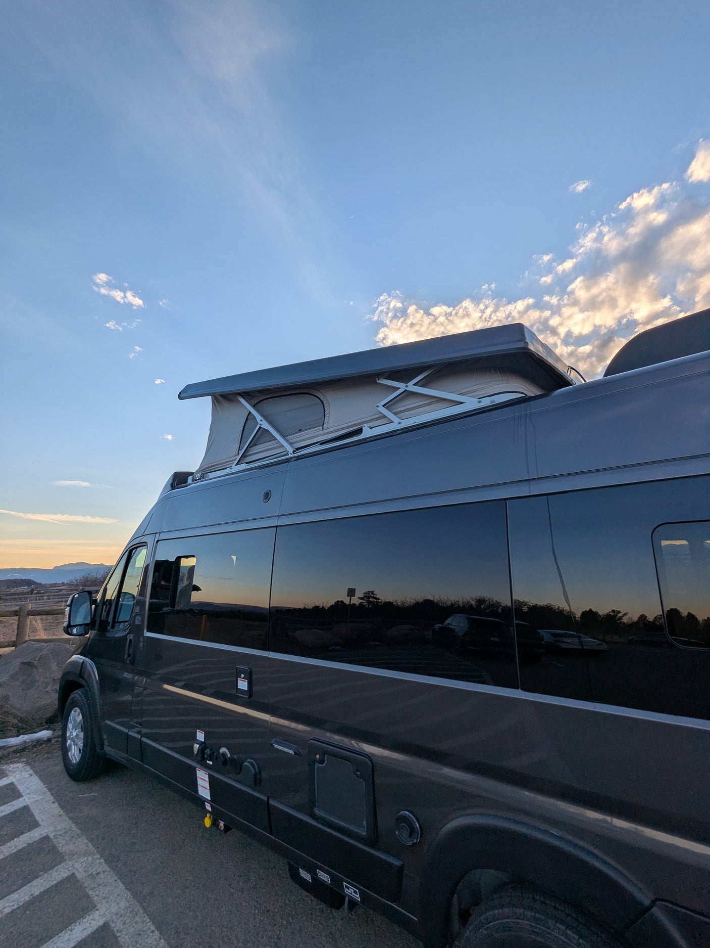 Dark grey RV rental with an extended pop-top tent, parked on asphalt. Sunset sky reflects in windows.