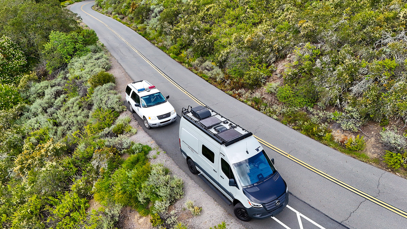 Modern Sprinter van rv rental, equipped with solar panels, follows a white SUV on a winding, nature-filled road.