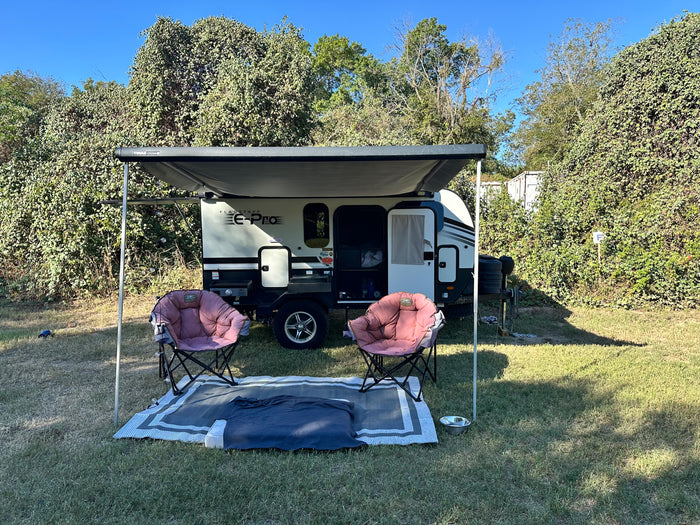 A compact RV rental with its awning extended, featuring two pink camping chairs and a mat on a grassy site, surrounded by lush trees.