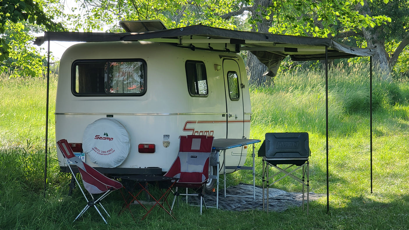 This cozy Scamp travel trailer, perfect for an RV rental, is set up at a shaded campsite with an awning, chairs, and grill.