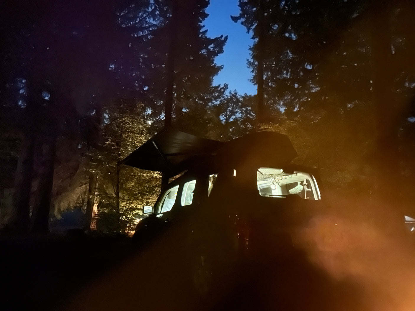 Cozy night camping with an RV rental, its awning extended, interior lit, amidst trees and campfire glow.
