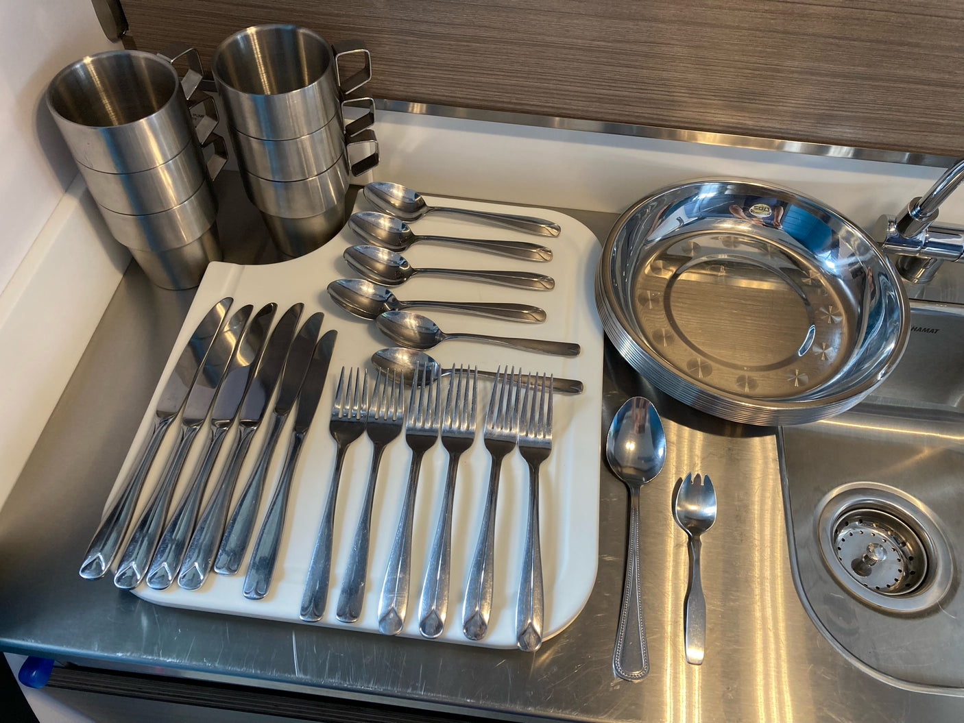 Compact RV rental kitchen features stainless steel cups, plates, and cutlery by the sink.