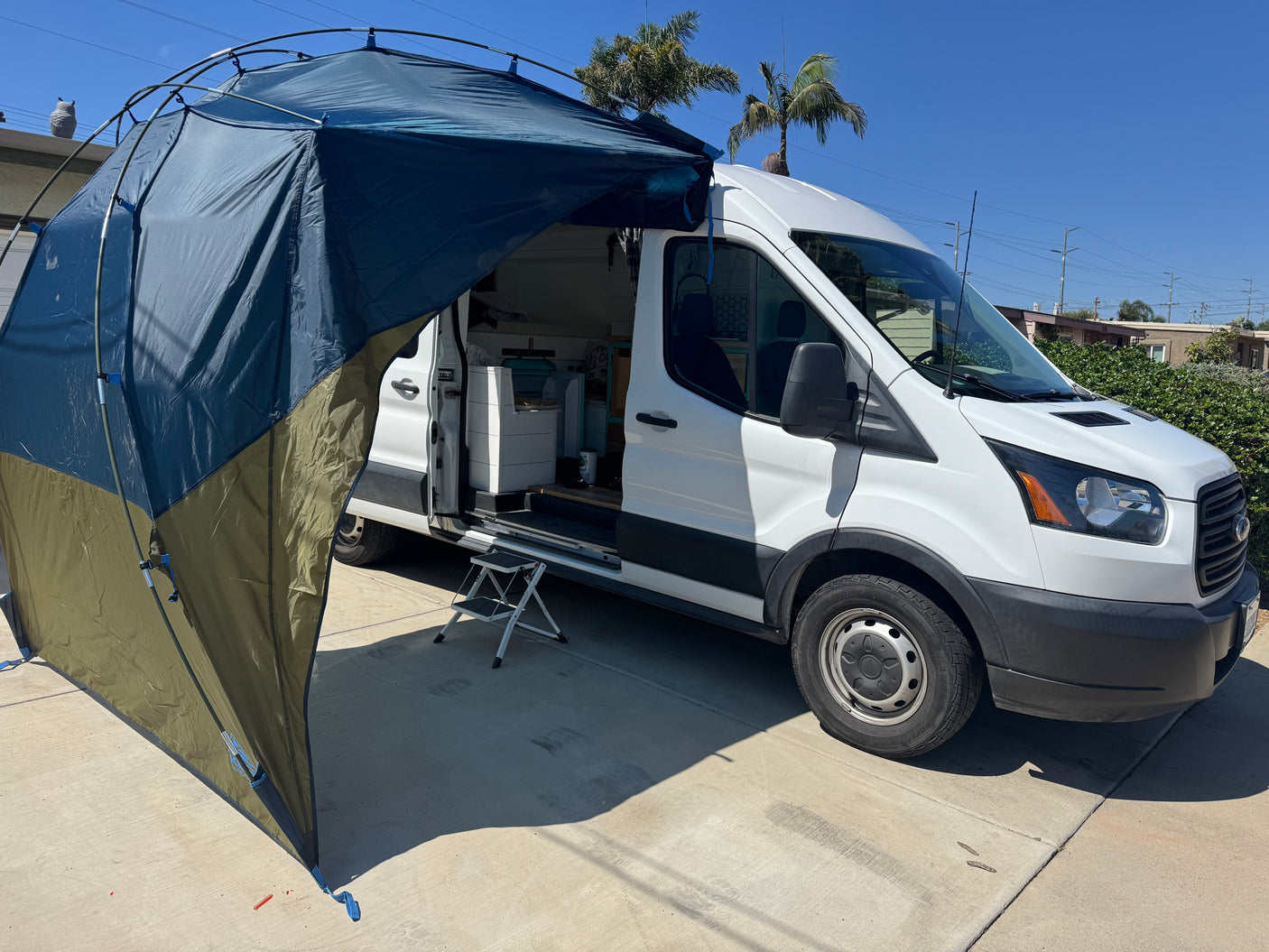 This white RV rental camper van features an attached privacy awning creating an outdoor living space. A step stool is by the open door.