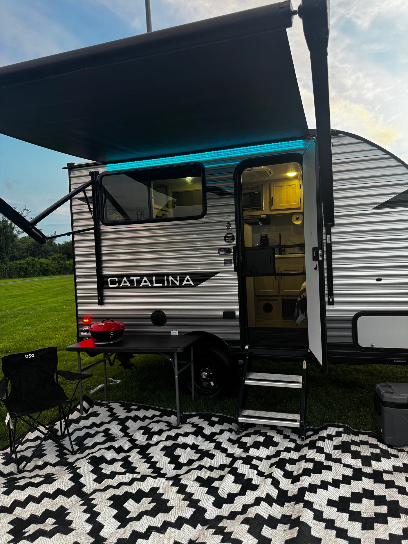 Evening at a Catalina RV rental: Awning with blue lights, steps, grill, chair, and patterned outdoor mat.