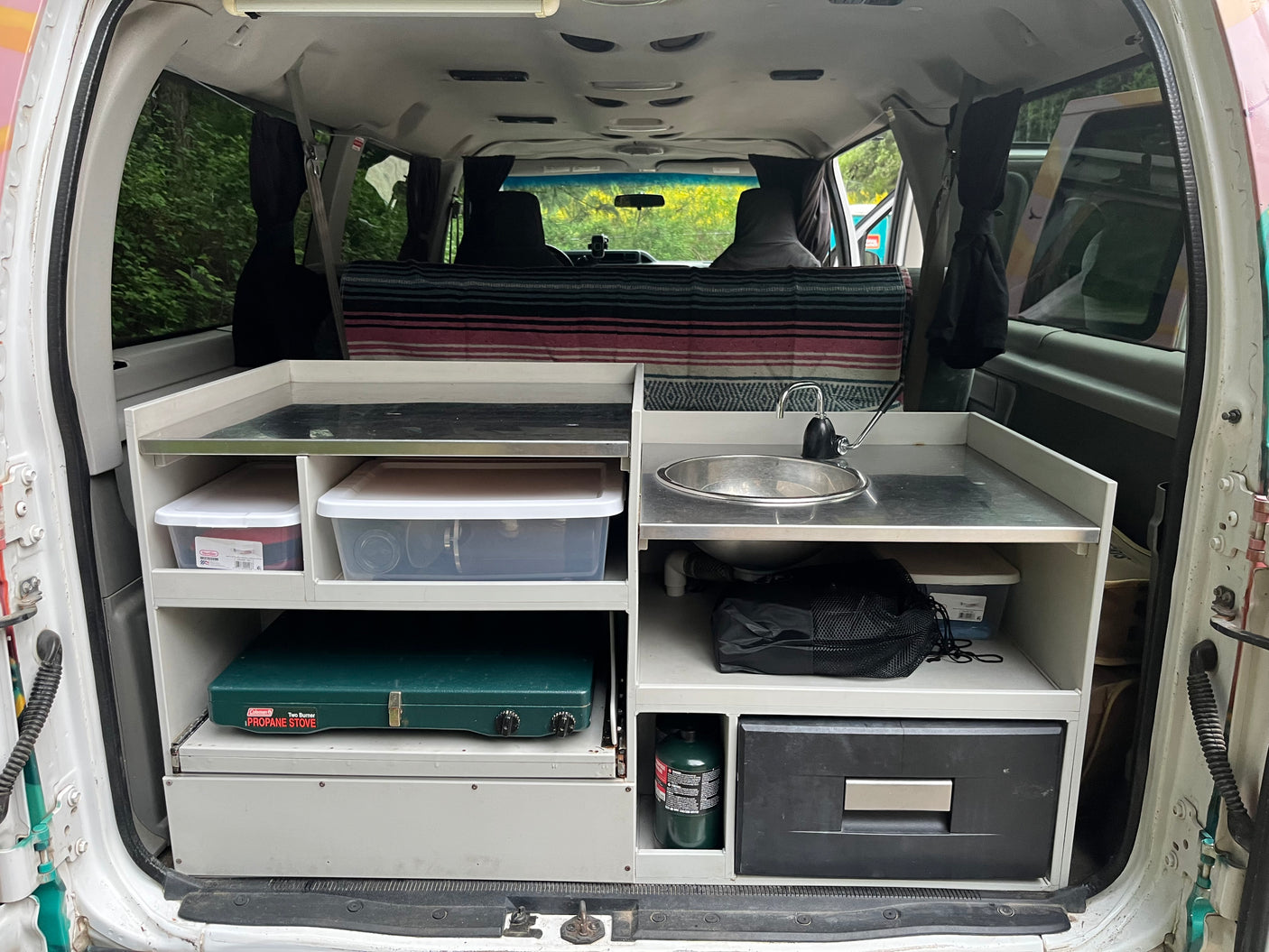 This rv rental features a compact camper van kitchen with a sink, stove, fridge, and storage bins for adventure.