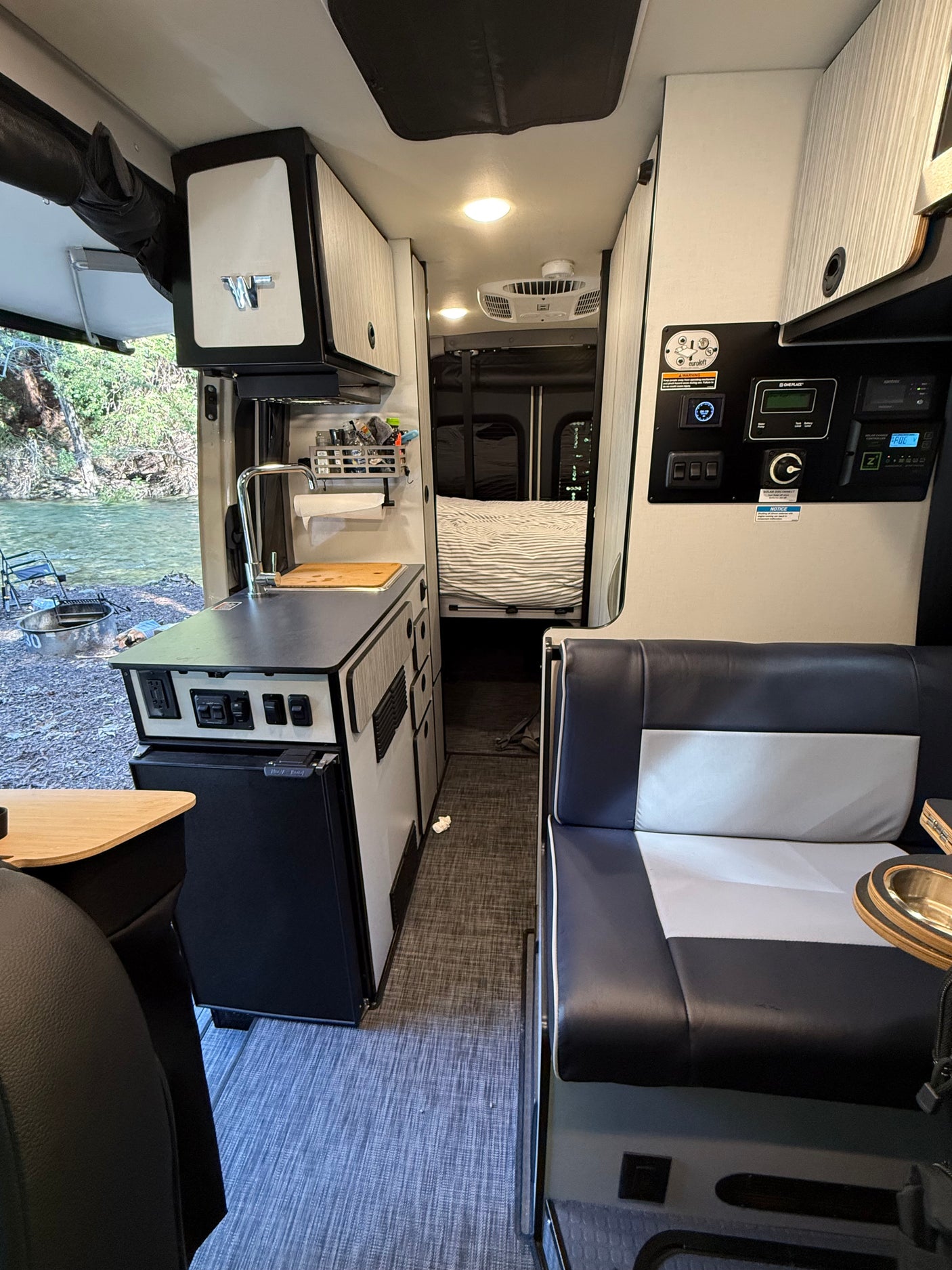 RV rental camper van interior featuring a kitchenette, dinette, and bed, with a scenic river view outside.