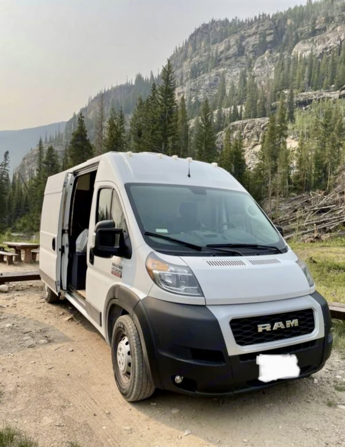 surrounded by scenic mountains.|White camper van RV rental parked by a scenic mountain lake. Perfect for adventure travel.|An RV rental's outdoor shower in action: A hand holds a nozzle spraying water from the rear of the white camper van.|This cozy RV rental features a comfortable van conversion interior with a bed