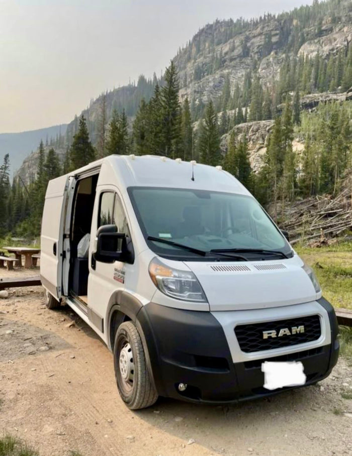 surrounded by scenic mountains.|White camper van RV rental parked by a scenic mountain lake. Perfect for adventure travel.|An RV rental's outdoor shower in action: A hand holds a nozzle spraying water from the rear of the white camper van.|This cozy RV rental features a comfortable van conversion interior with a bed