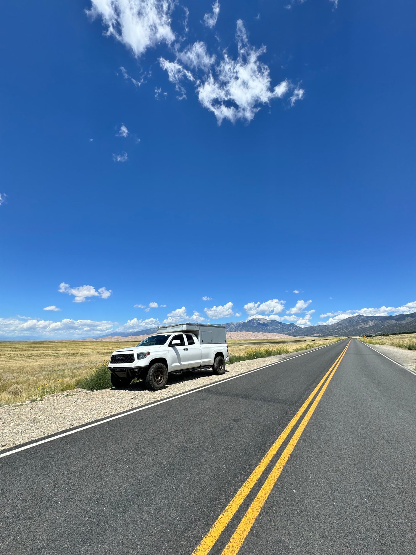 White pickup truck with camper, ready for an rv rental road trip, by golden fields, mountains & blue sky.