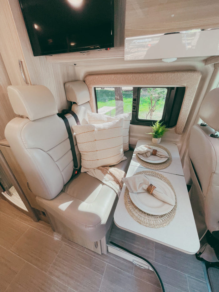 Inside a modern RV rental: a bright living area with dining table for two, comfortable seats, and a TV.