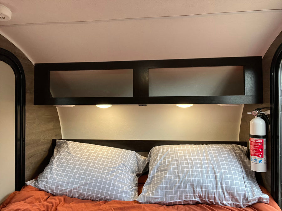 RV rental interior showing a bed with pillows, overhead cabinets, reading lights, and a visible fire extinguisher for safety.