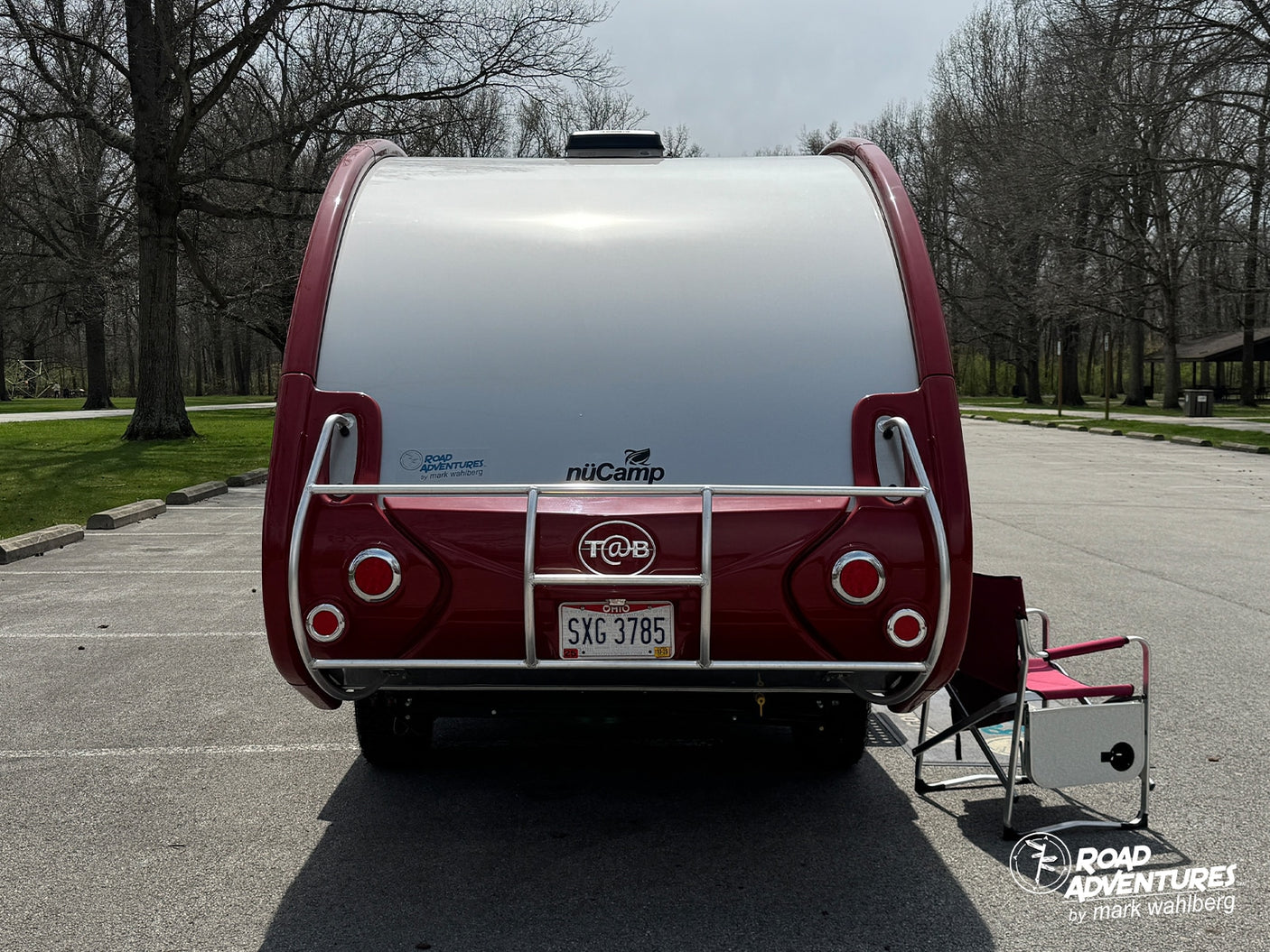 Rear view of a red and white nüCamp T@B travel trailer, an ideal rv rental for park adventures. A camping chair sits nearby.