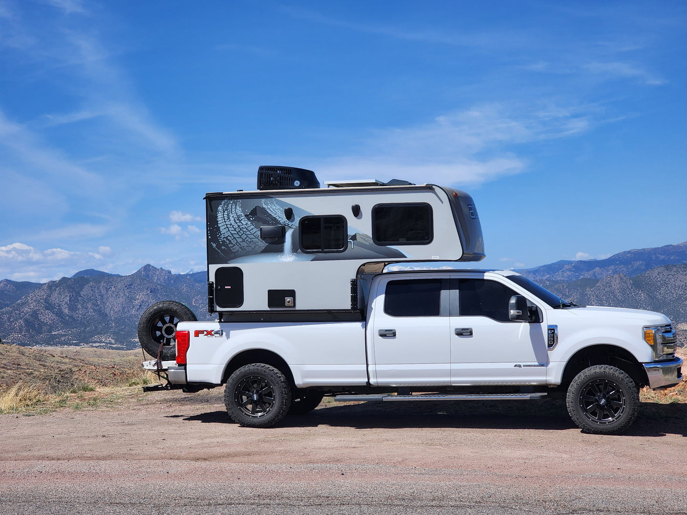 White Ford truck with a large, grey truck bed RV camper, featuring a tire tread graphic, parked on a dirt road overlooking mountains. An ideal RV rental.