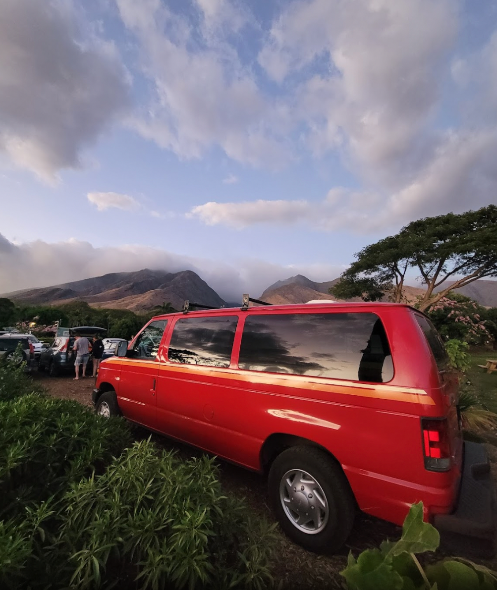 Red RV rental van amidst lush plants, with majestic cloud-covered mountains & dynamic sky in the background, perfect for road trips.