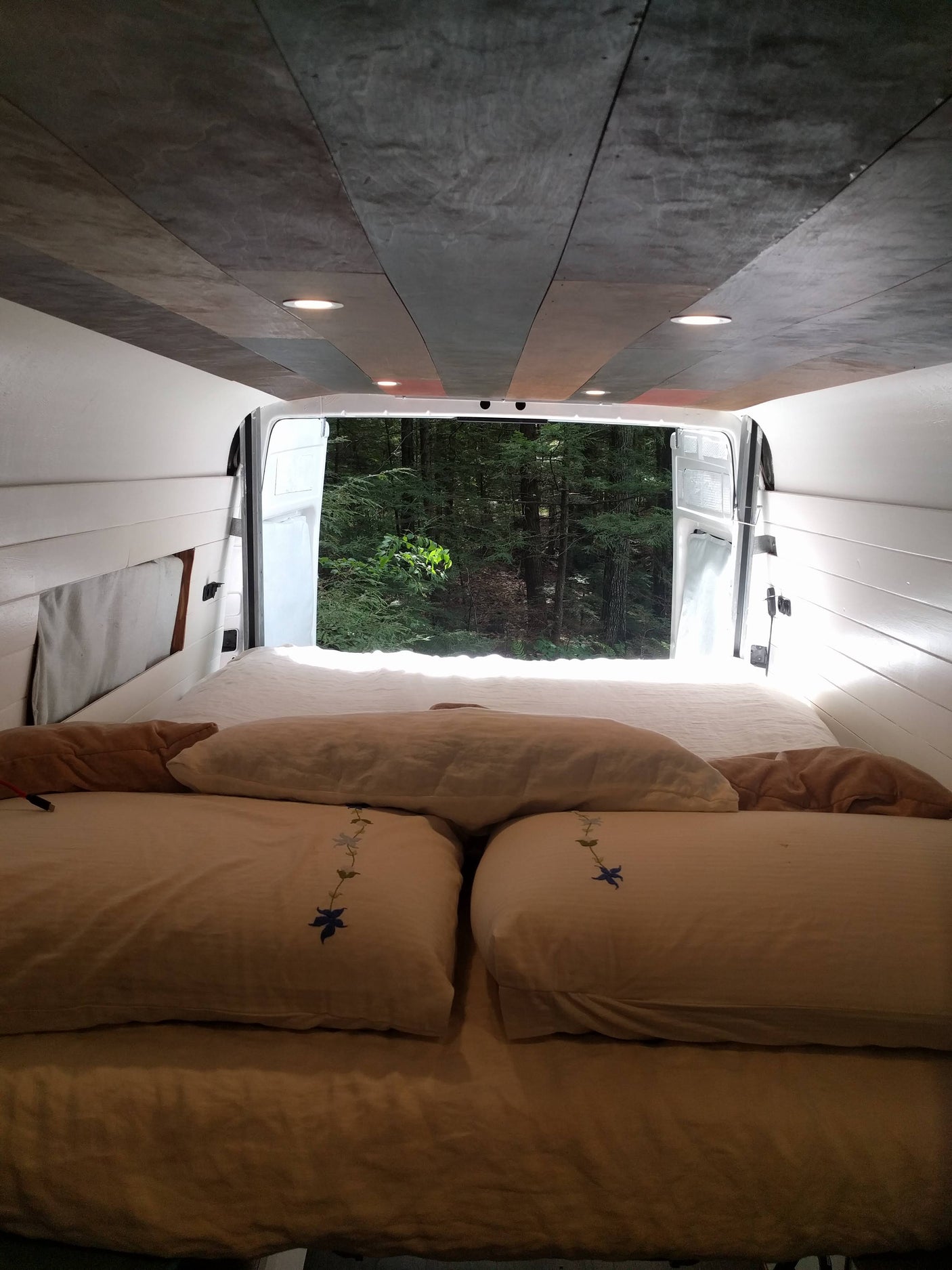 Relax in this cozy RV rental's bed, looking out at a tranquil forest.
