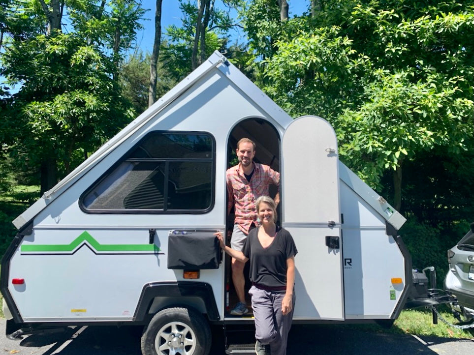 A smiling man and woman stand by their white A-frame RV rental, ready for outdoor adventure.