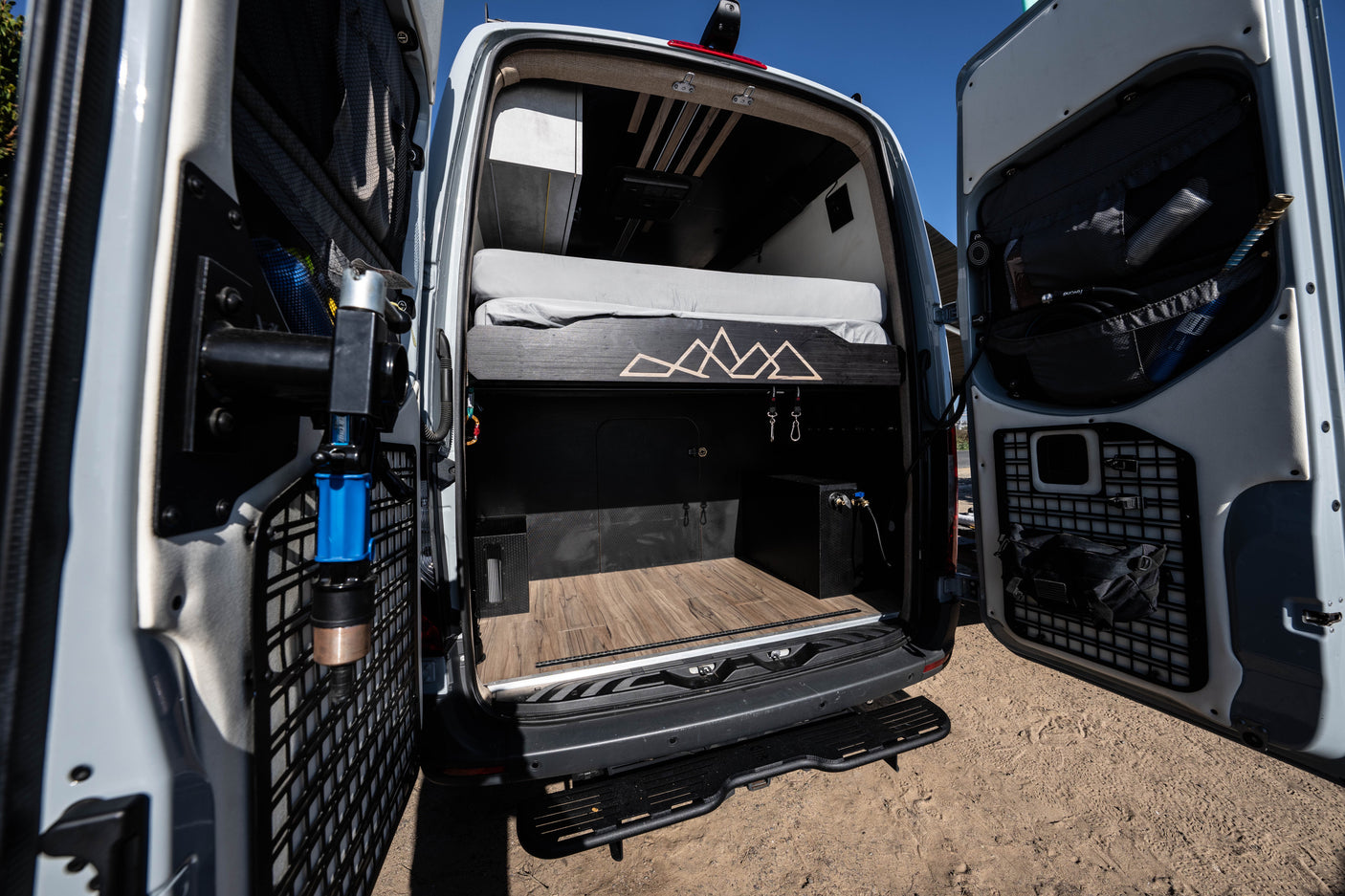 Inside a custom rv rental: rear view of a camper van's organized storage area with a bed, wood floor, and door panels.
