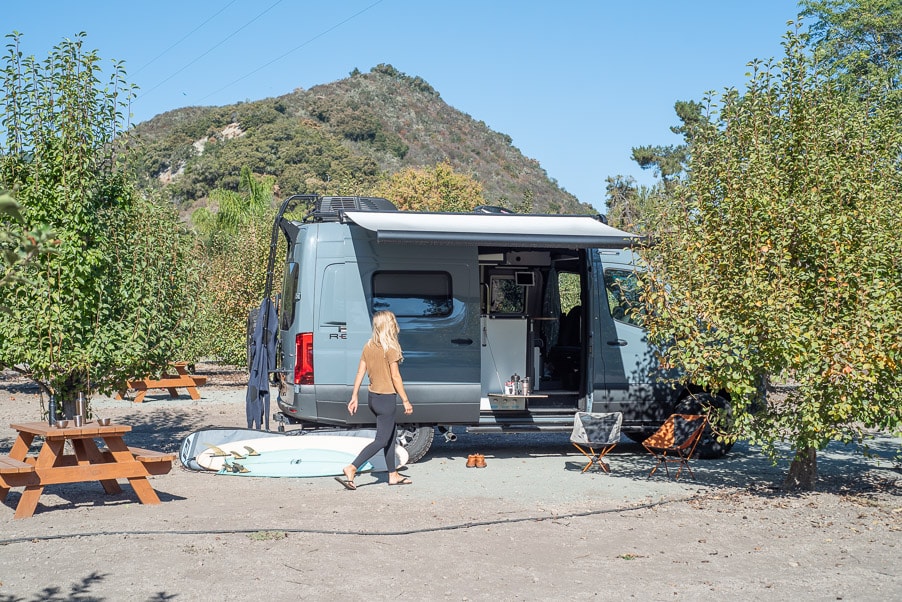 Grey RV rental camper van parked with open side. A woman walks towards it. Surfboard, chairs & picnic tables nearby. — photo 5