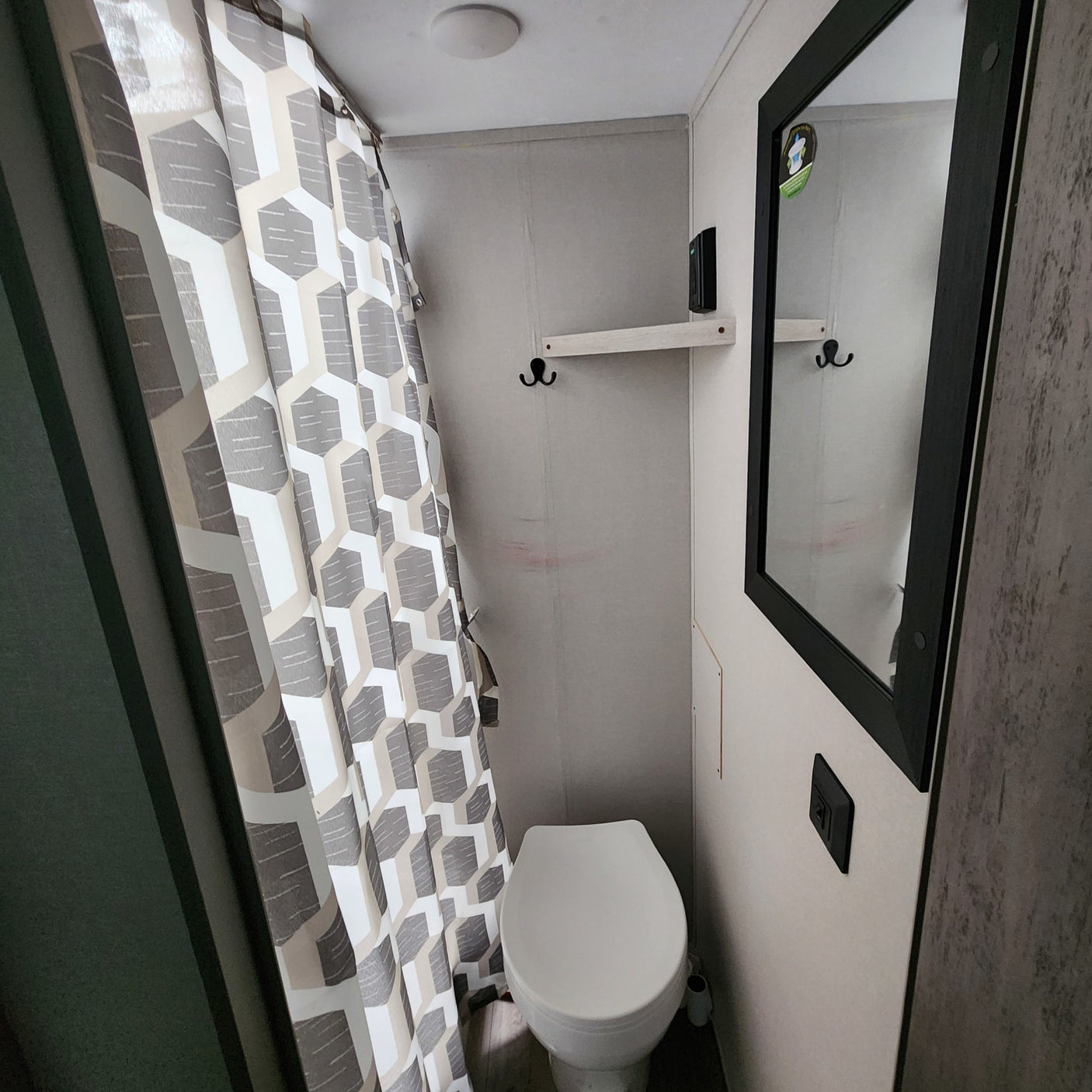 Compact RV rental bathroom features a white toilet, geometric gray/white shower curtain, mirror, and small shelf with hooks.