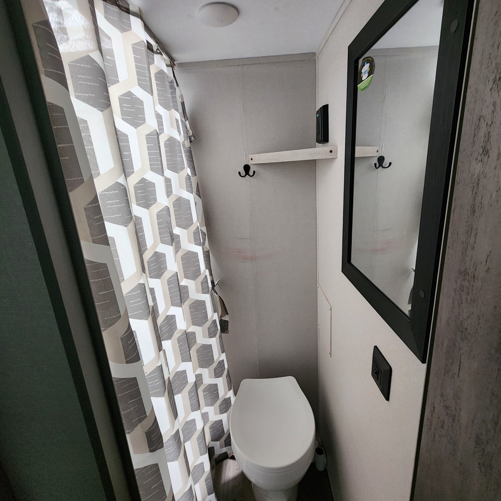 Compact RV rental bathroom features a white toilet, geometric gray/white shower curtain, mirror, and small shelf with hooks.