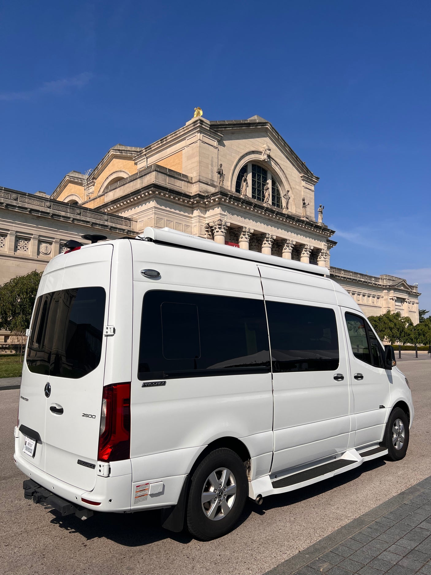 White Mercedes Sprinter RV rental, a luxury camper van, stands before a grand, classical-style building under a clear blue sky.