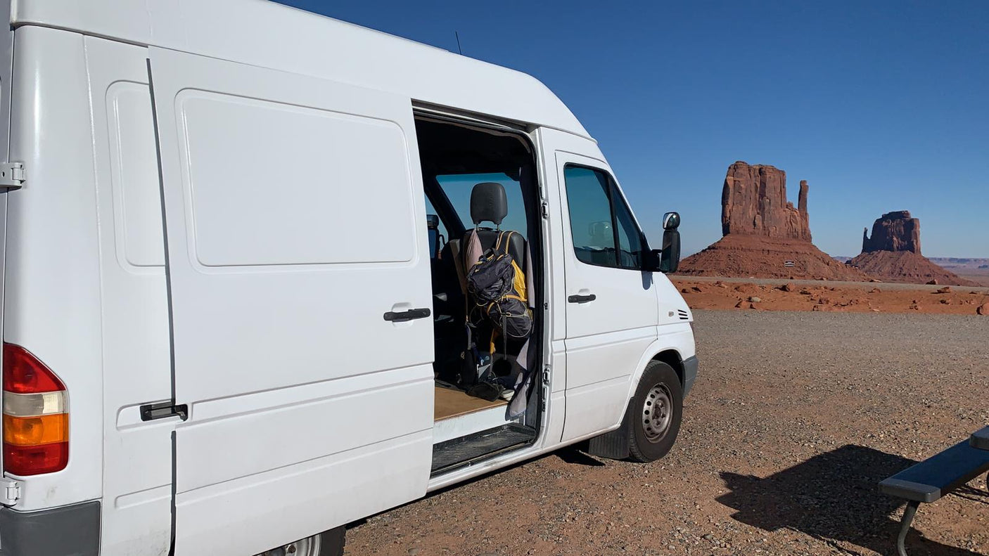A white rv rental van, side door open, parked in a desert lot. Iconic red rock formations of Monument Valley rise under a blue sky.