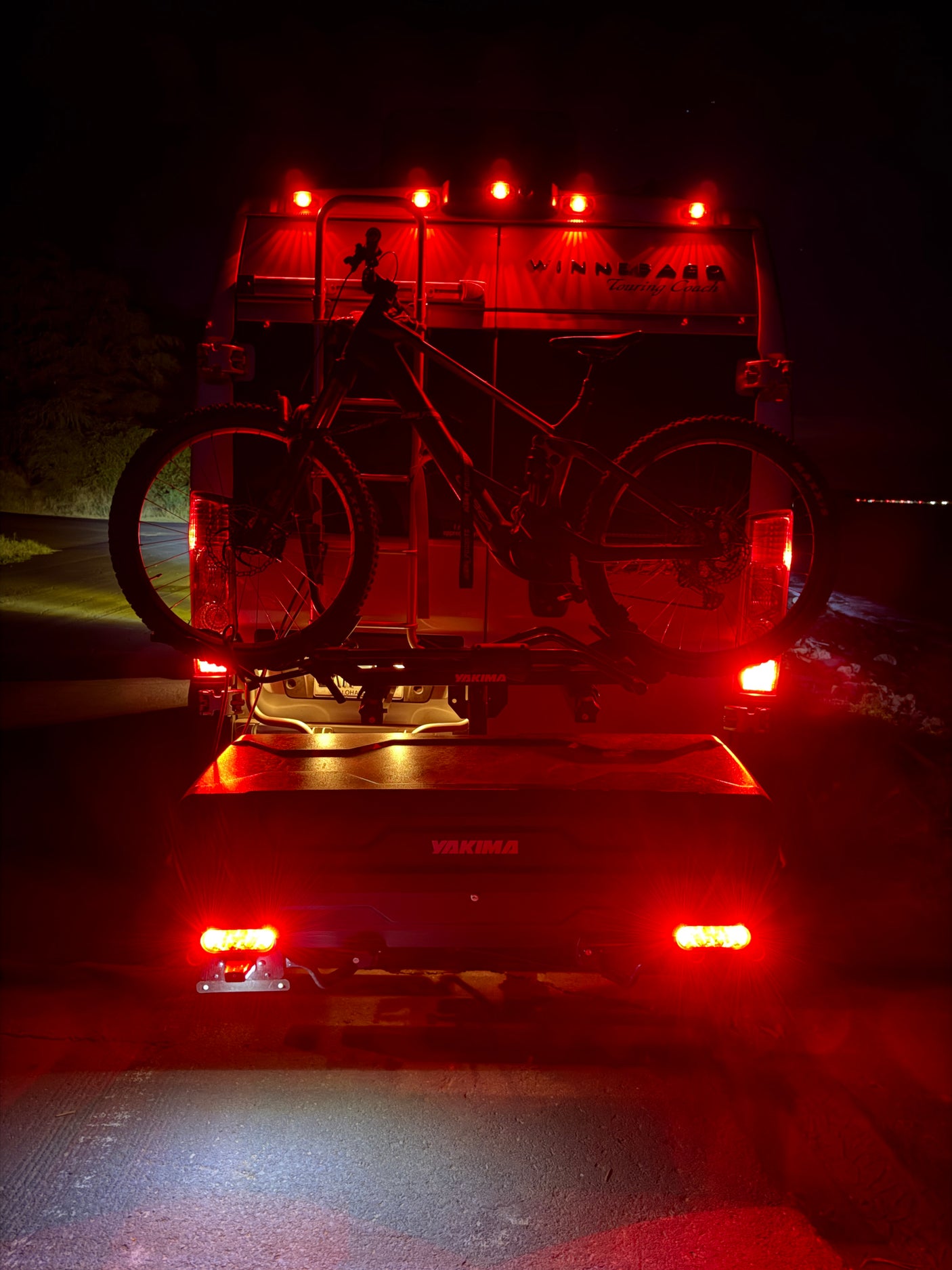 An RV rental at night, with a mountain bike on a Yakima rack and cargo box attached, illuminated by bright red taillights.