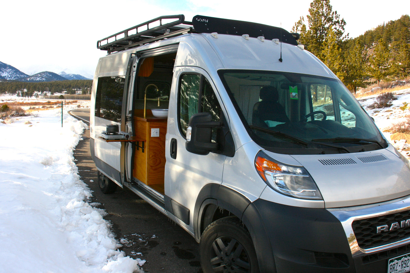 Silver RV rental van, with side door open to reveal a compact kitchen, parked by a snowy road amidst mountains.