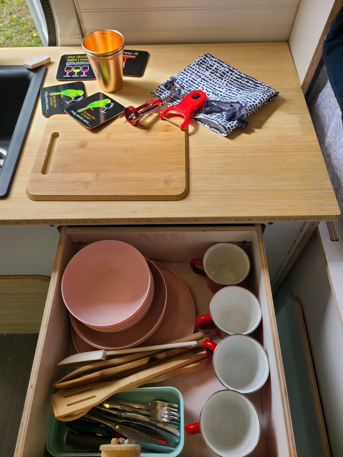 RV rental's compact kitchen boasts a bamboo counter, cutting board, and organized drawer with pink plates, mugs, & utensils.