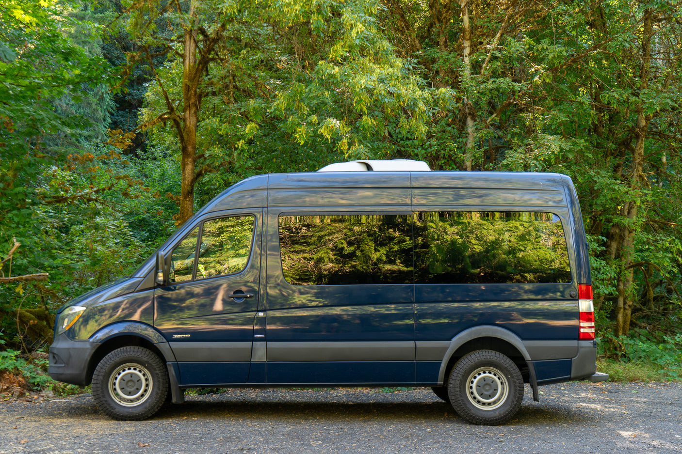 Dark blue Mercedes Sprinter RV rental parked in a lush green forest, ready for outdoor adventure.